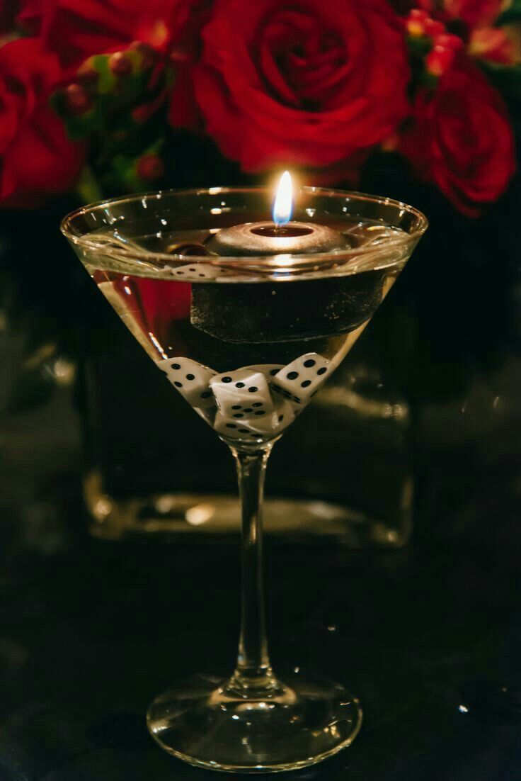 a candle is lit in a martini glass with dice on the rim and red roses behind it