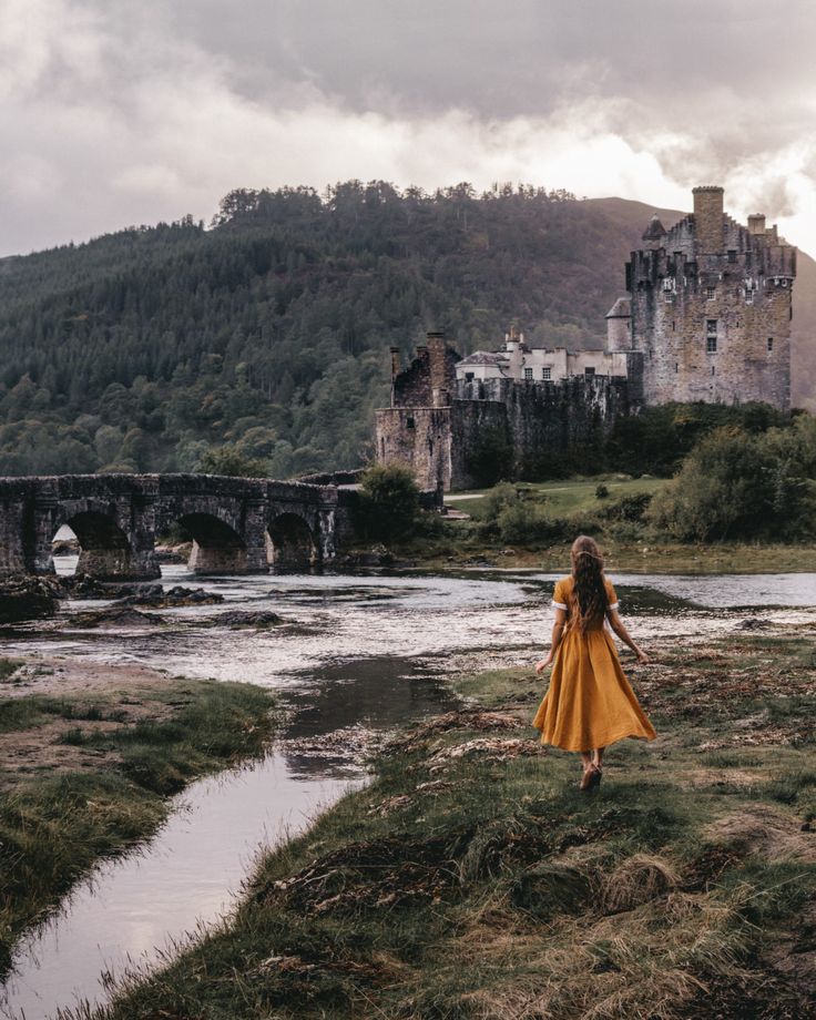 a girl in a yellow dress is standing on the grass near a river and castle