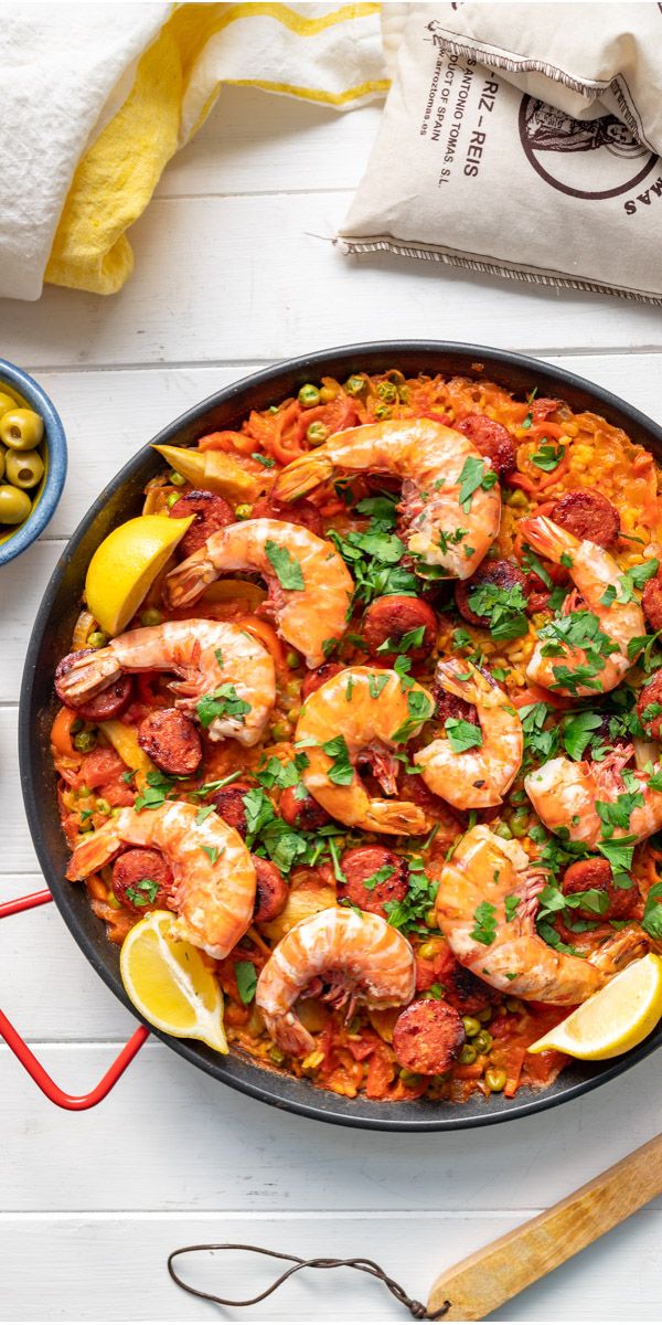 a skillet filled with shrimp and rice next to olives, lemon wedges and tomatoes