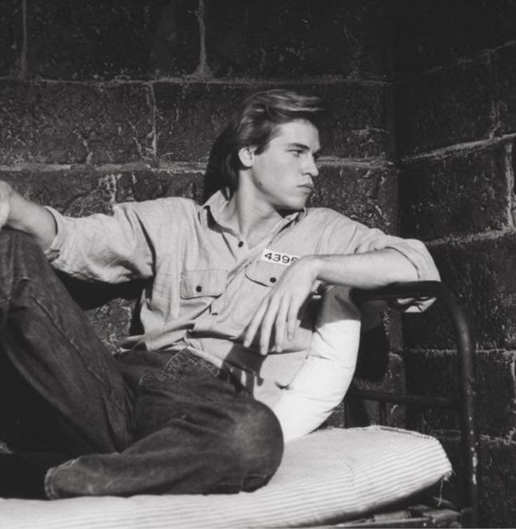 a black and white photo of a man sitting on a bed in front of a brick wall