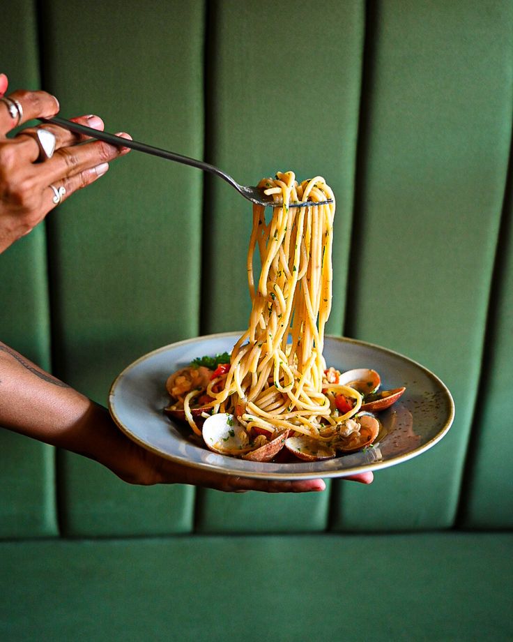 a person is holding a plate with noodles and vegetables on it, while the other hand holds a fork