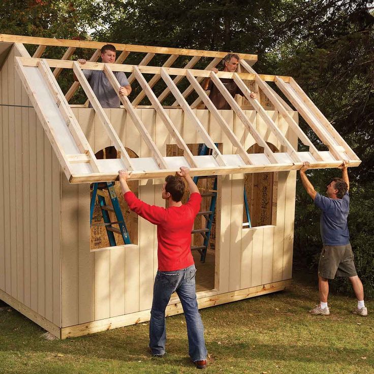 two men are building a small house out of wood and sidings, while another man holds up the roof