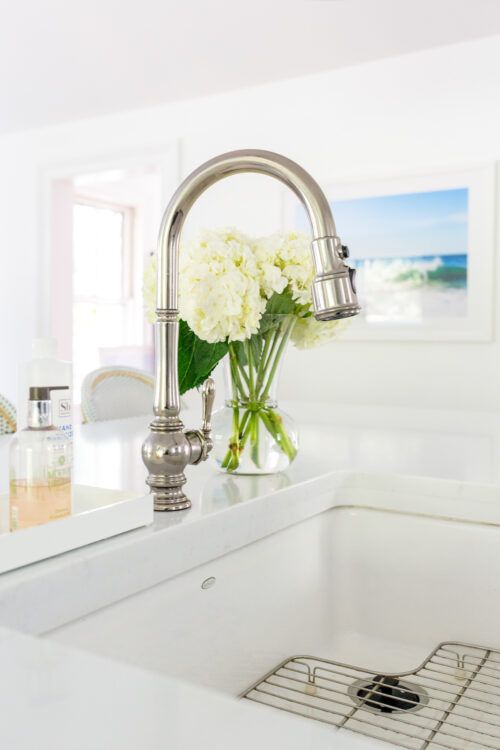 a white kitchen sink sitting under a window next to a faucet with flowers in it