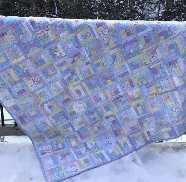 a quilt hanging on a fence in the snow with trees in the backgroud