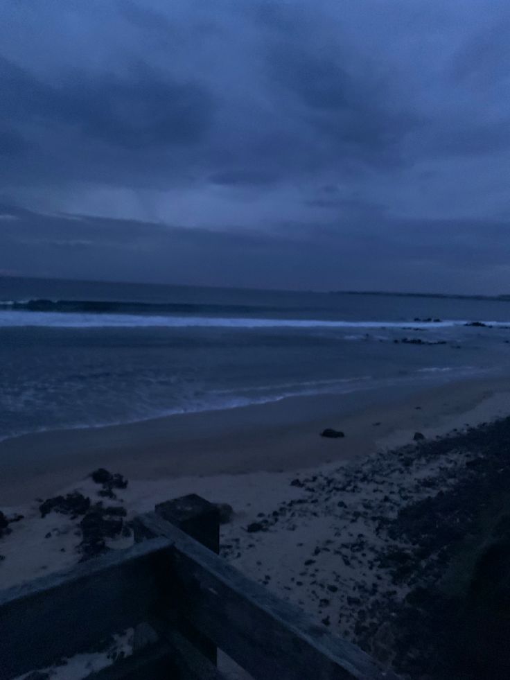 an empty beach at night with the ocean in the distance and dark clouds above it