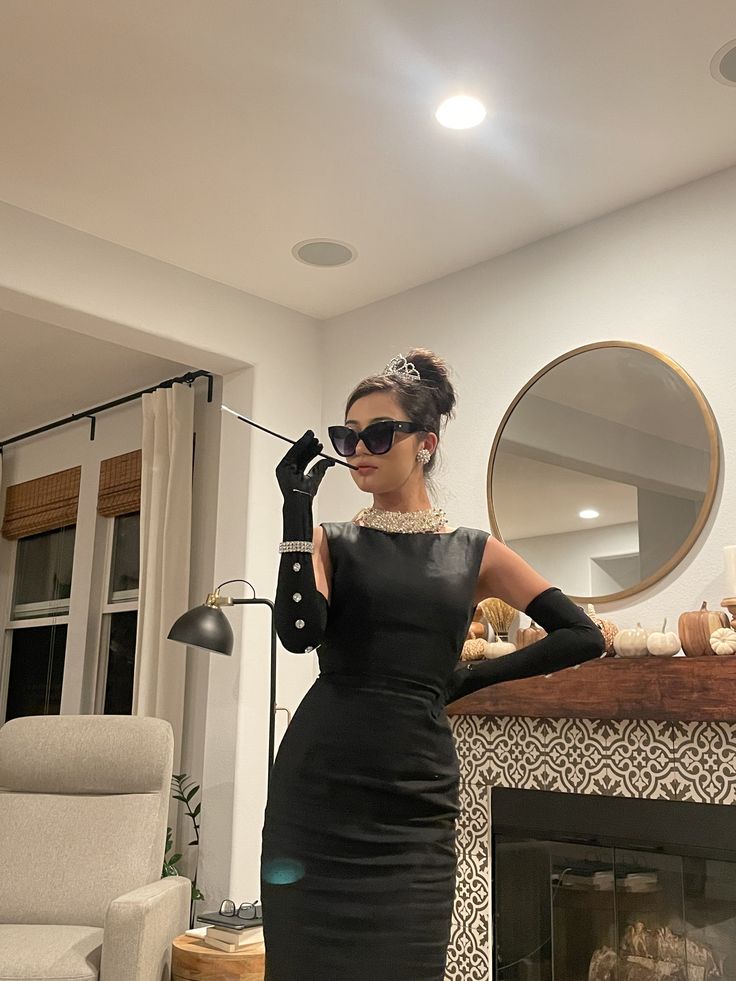 a woman in a black dress is standing near a fireplace and holding a phone up to her ear