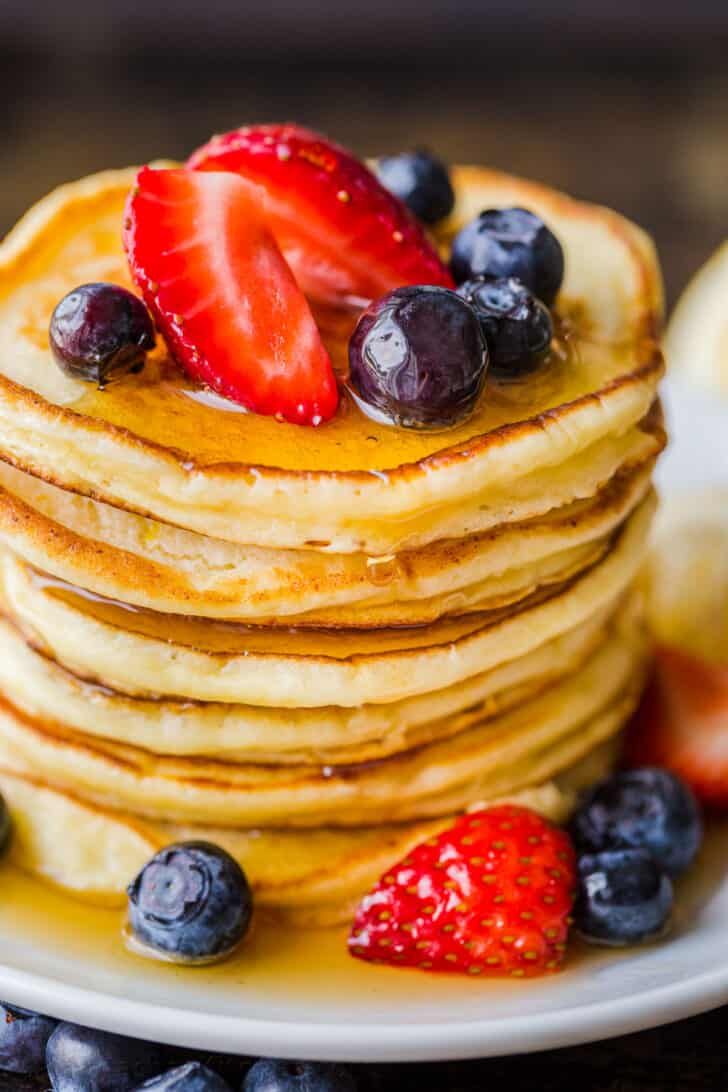 a stack of pancakes topped with strawberries and blueberries being drizzled with syrup