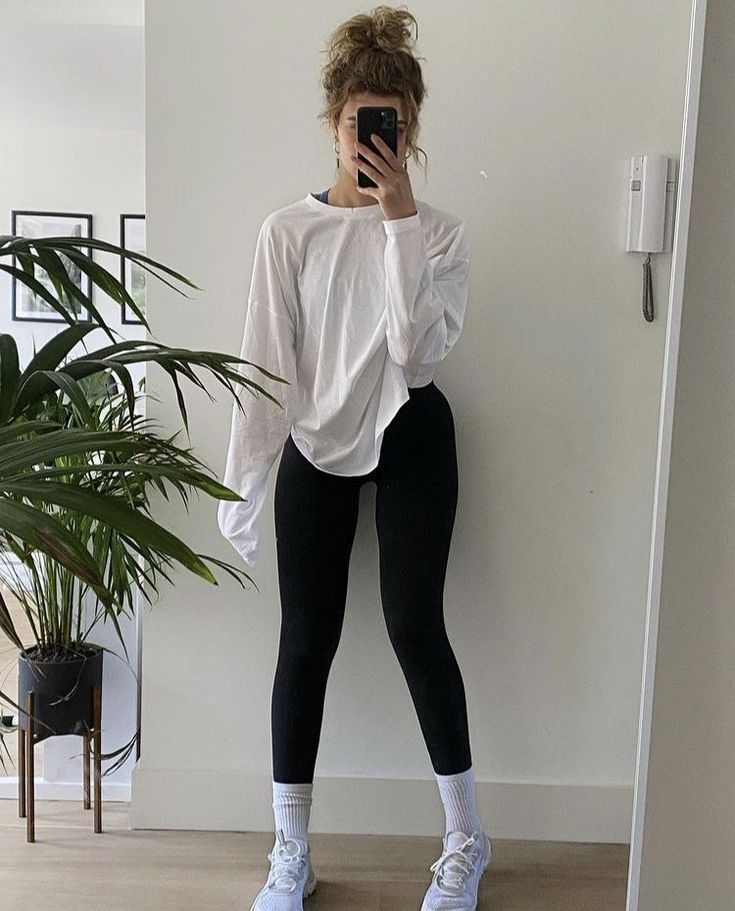 a woman in white shirt and black leggings taking a selfie with her cell phone