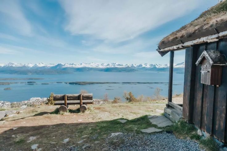 an outhouse on the side of a hill overlooking water and snow capped mountains in the distance