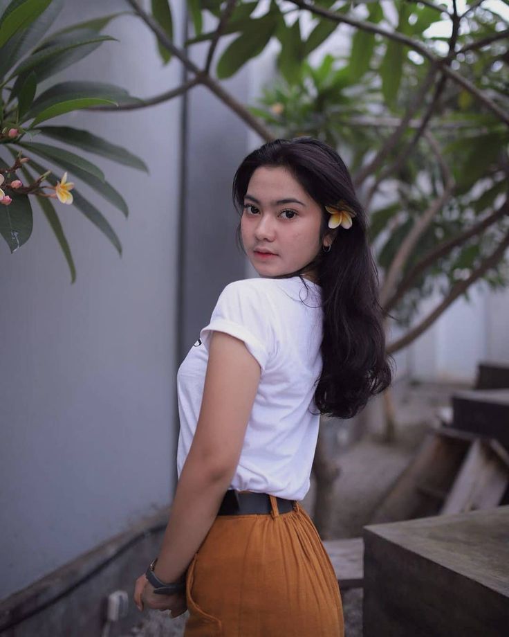 a woman standing next to a tree wearing a white shirt and brown skirt with flowers in her hair