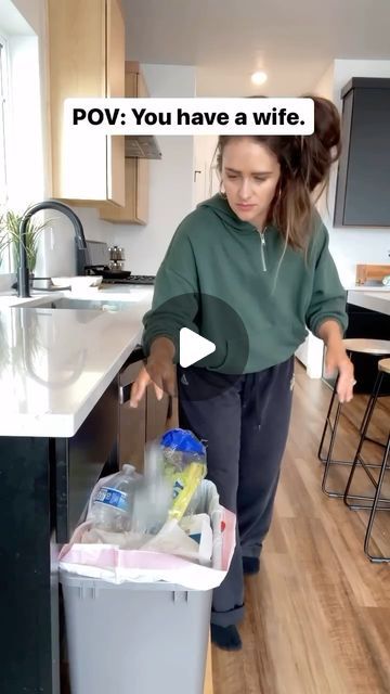 a woman standing in front of a kitchen counter next to a trash can filled with plastic bottles