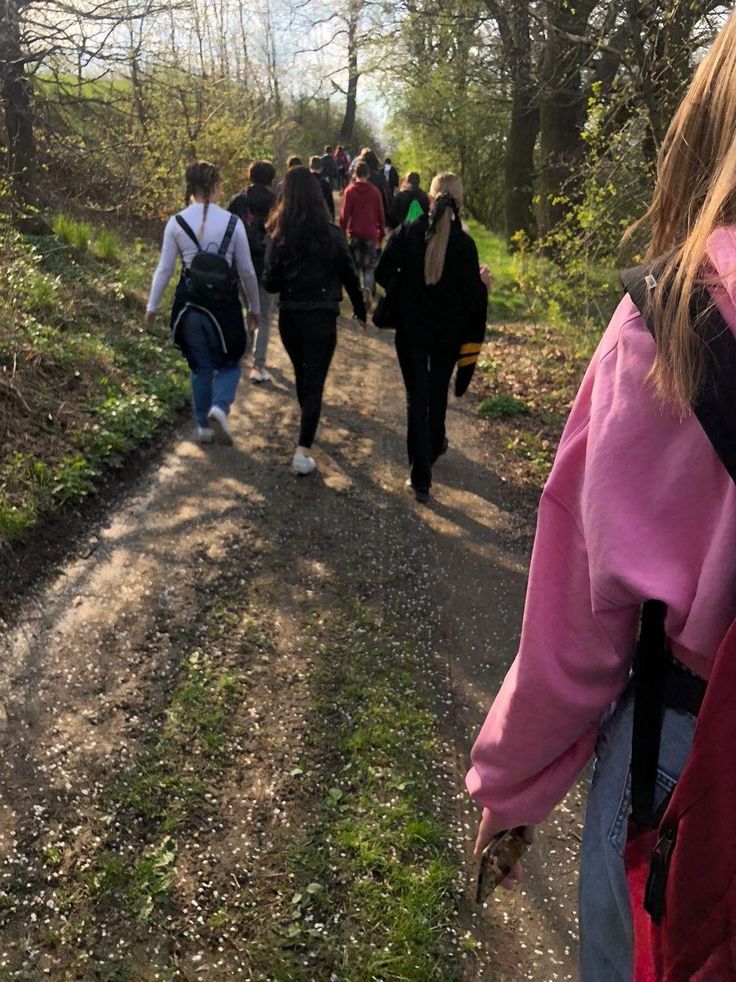 a group of people walking down a dirt road in the woods on a sunny day