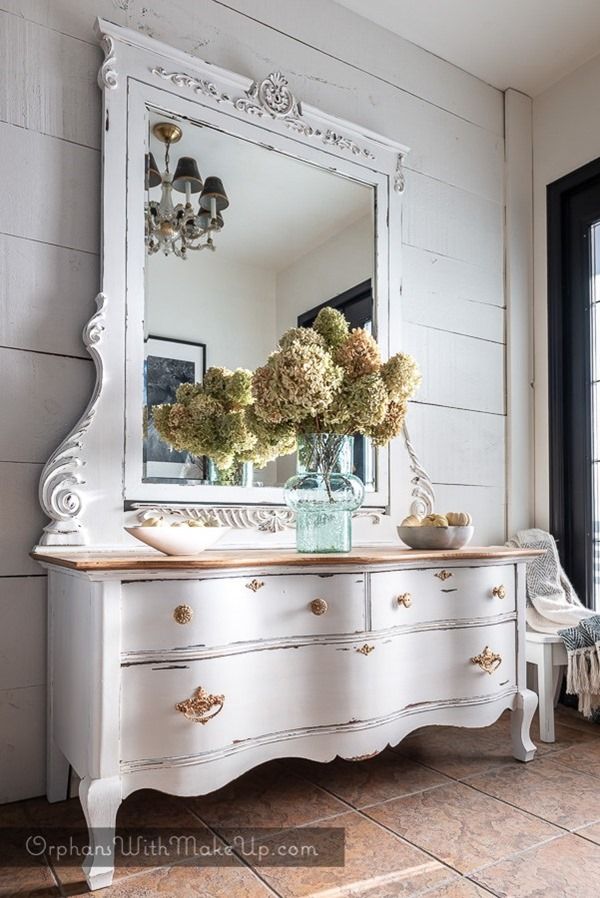 a white dresser with flowers in a vase on top and a large mirror above it