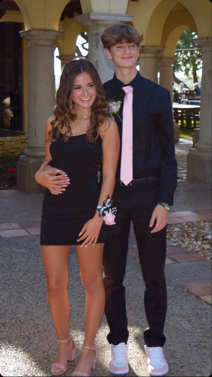 a young man standing next to a woman in a short black dress and pink tie