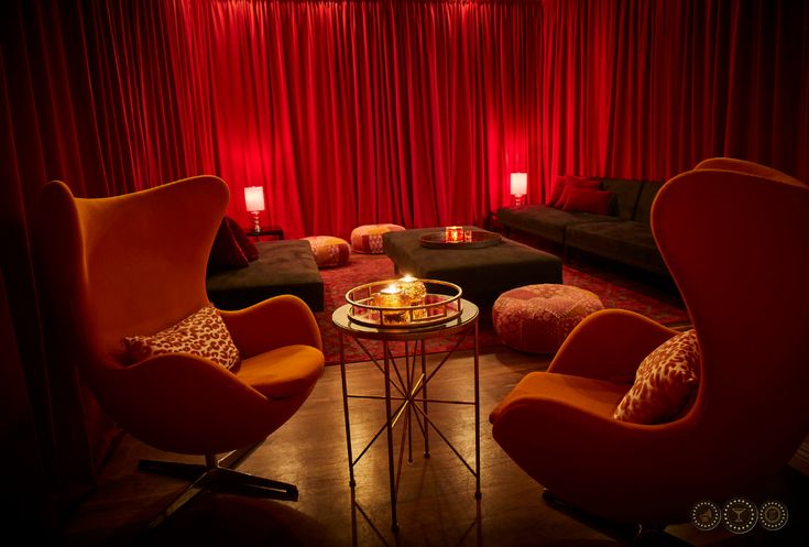 a living room filled with furniture and red drapes on the windows behind it is a coffee table surrounded by two orange chairs