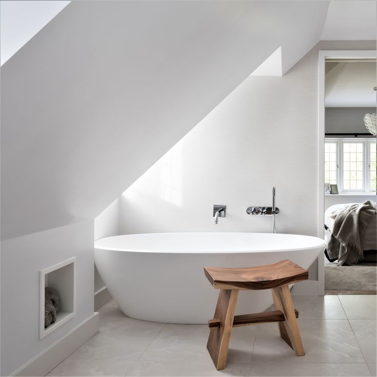 a large white bathtub sitting under a slanted ceiling next to a wooden stool