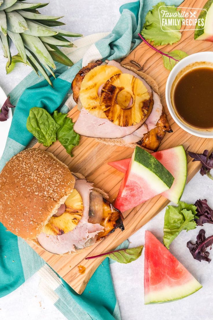 two sandwiches with meat, pineapples and watermelon slices on a cutting board