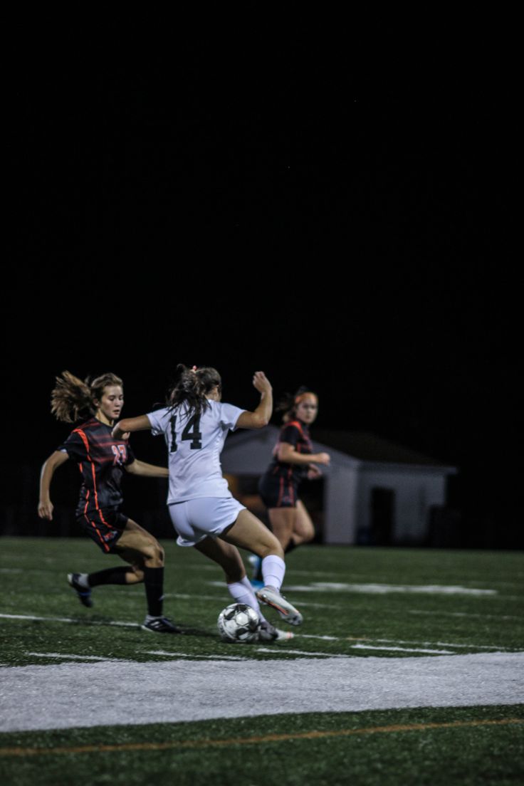 two girls playing soccer on a field at night with the ball in front of them