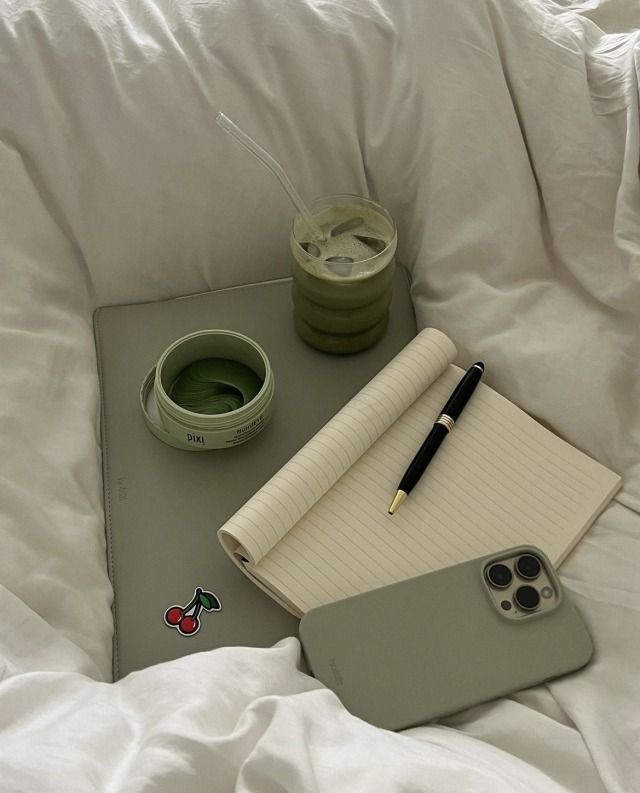 a notepad, pen and cup sitting on top of a bed with white sheets