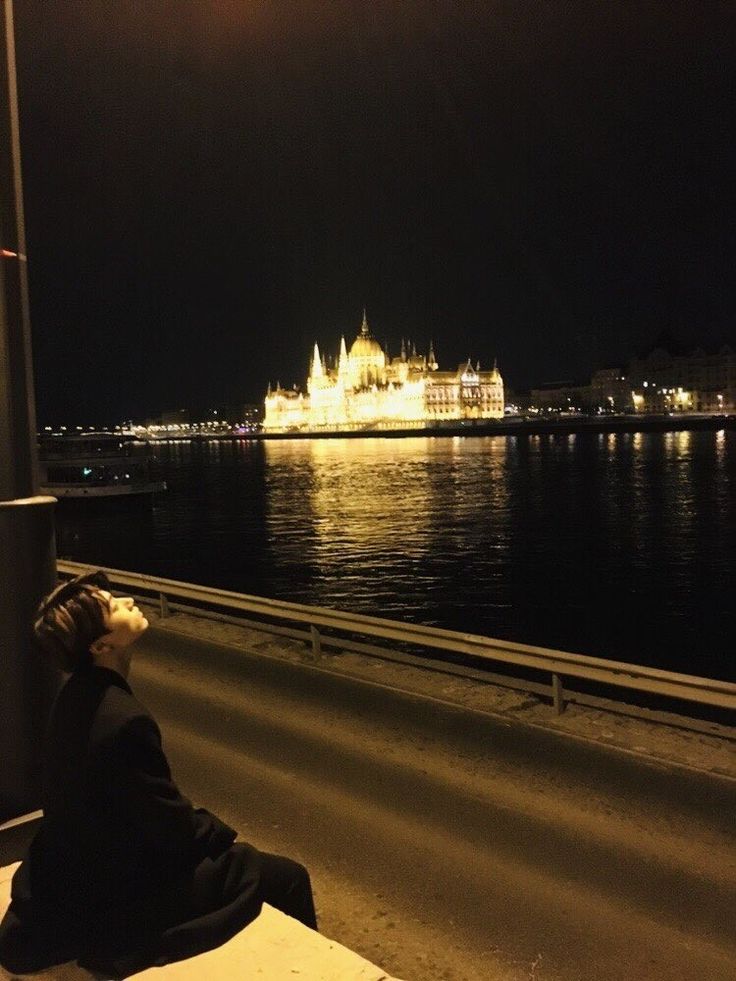 a person sitting on a bench next to the water at night with a castle in the background