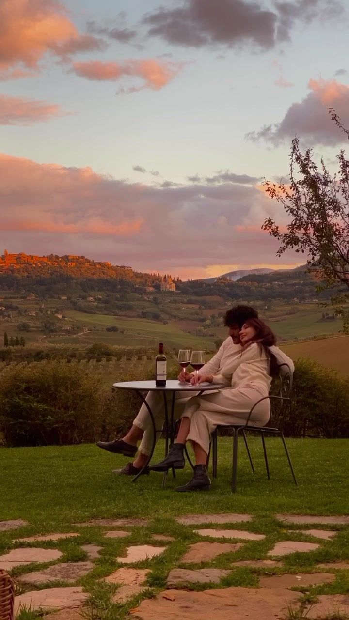 a woman sitting at a table with a bottle of wine in her hand and the sun setting behind her