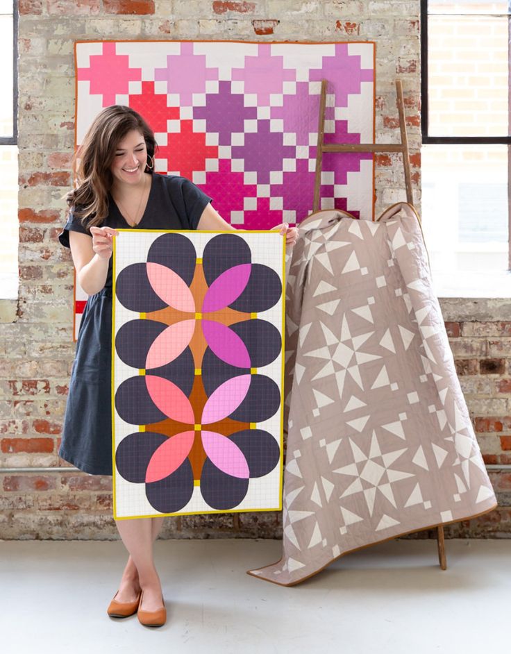 a woman holding up a large quilt next to a chair and another piece of art