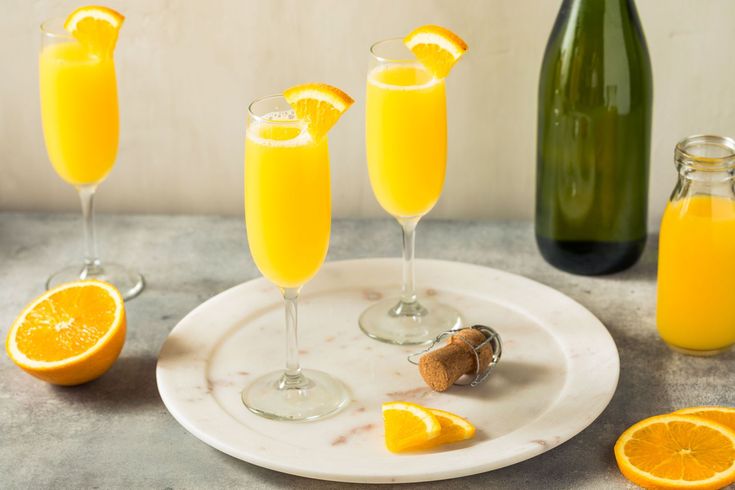 three glasses of orange juice on a plate next to two bottles and one glass with an orange slice