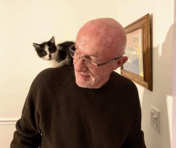 an old man with glasses and a cat sitting on his shoulder looking at him in the mirror