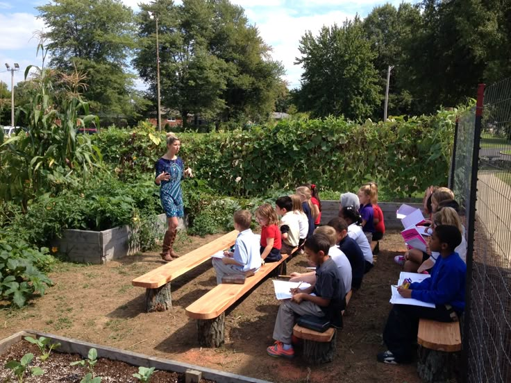 a group of children sitting on wooden benches in a garden with an adult standing next to them