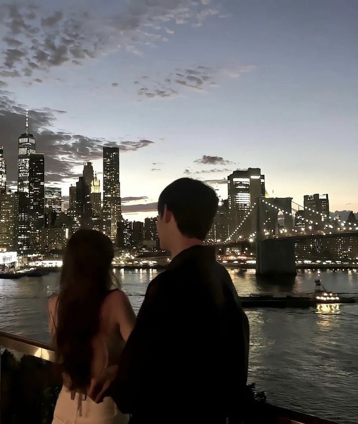 a man and woman looking out over the water at night time in new york city