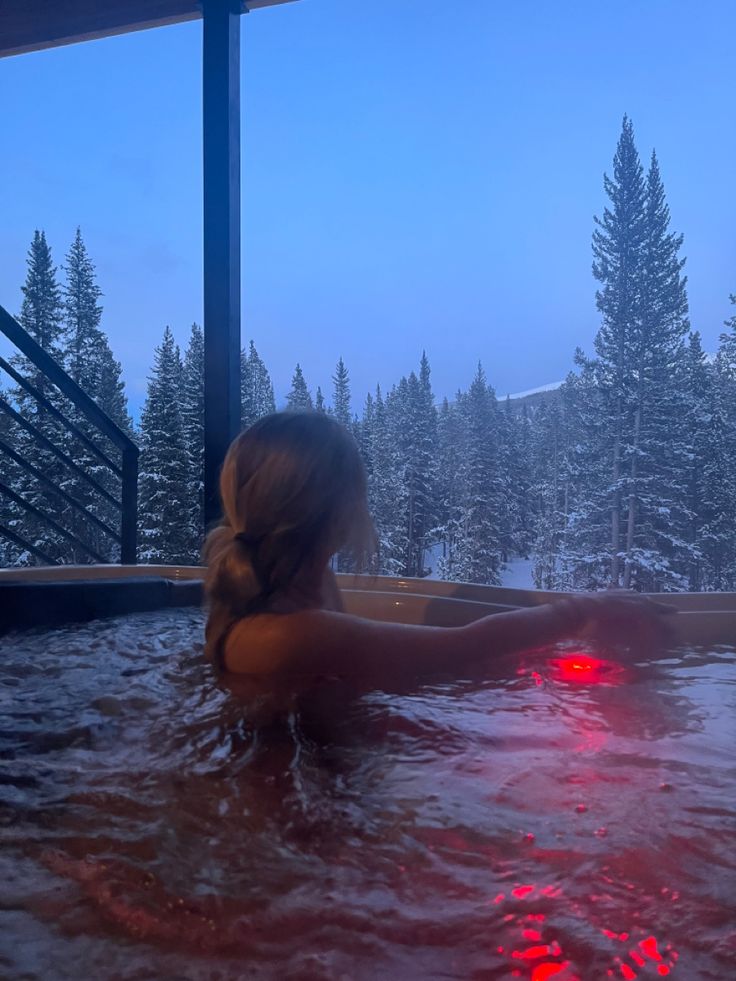 a woman sitting in a hot tub with red lights on it's side and snow covered trees behind her