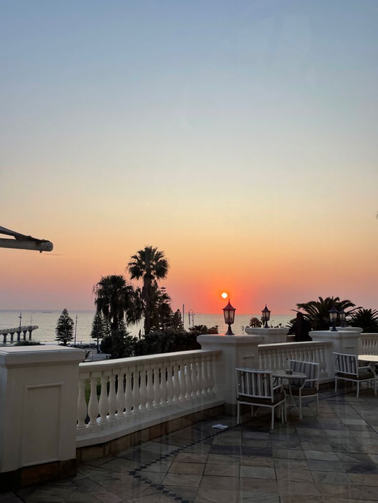 the sun is setting over the ocean with palm trees in the foreground and benches on the other side