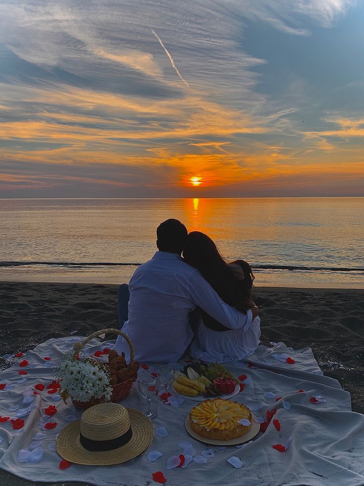 two people sitting on a blanket at the beach with food and drinks in front of them