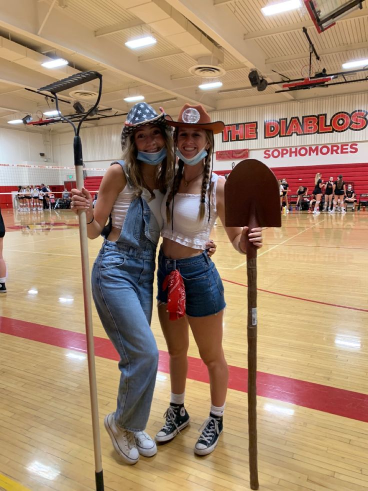 two women in overalls and hats pose for a photo on a basketball court with paddles