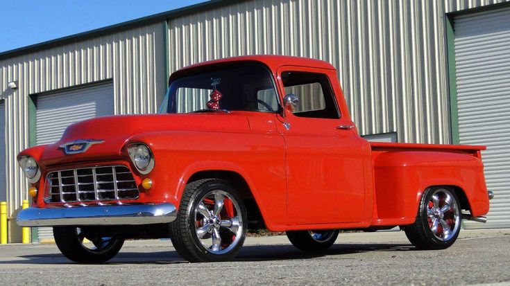 an old red truck is parked in front of a garage door and some metal buildings