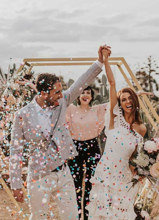 a newly married couple walk through confetti as they exit the ceremony with their arms in the air