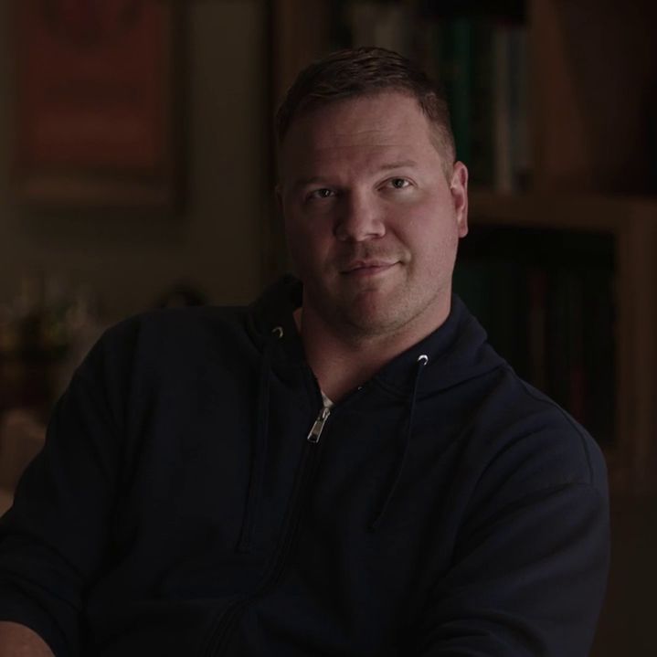 a man sitting in front of a bookshelf with his arms crossed and looking at the camera