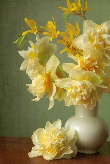 a white vase with yellow flowers in it on a wooden table next to a green wall