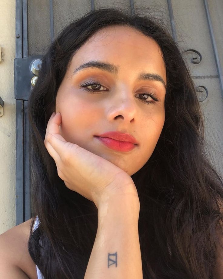 a woman with long black hair wearing a white tank top and holding her hand on her chin