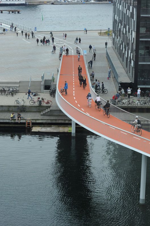 people are walking and riding bikes on an orange bridge over water with buildings in the background