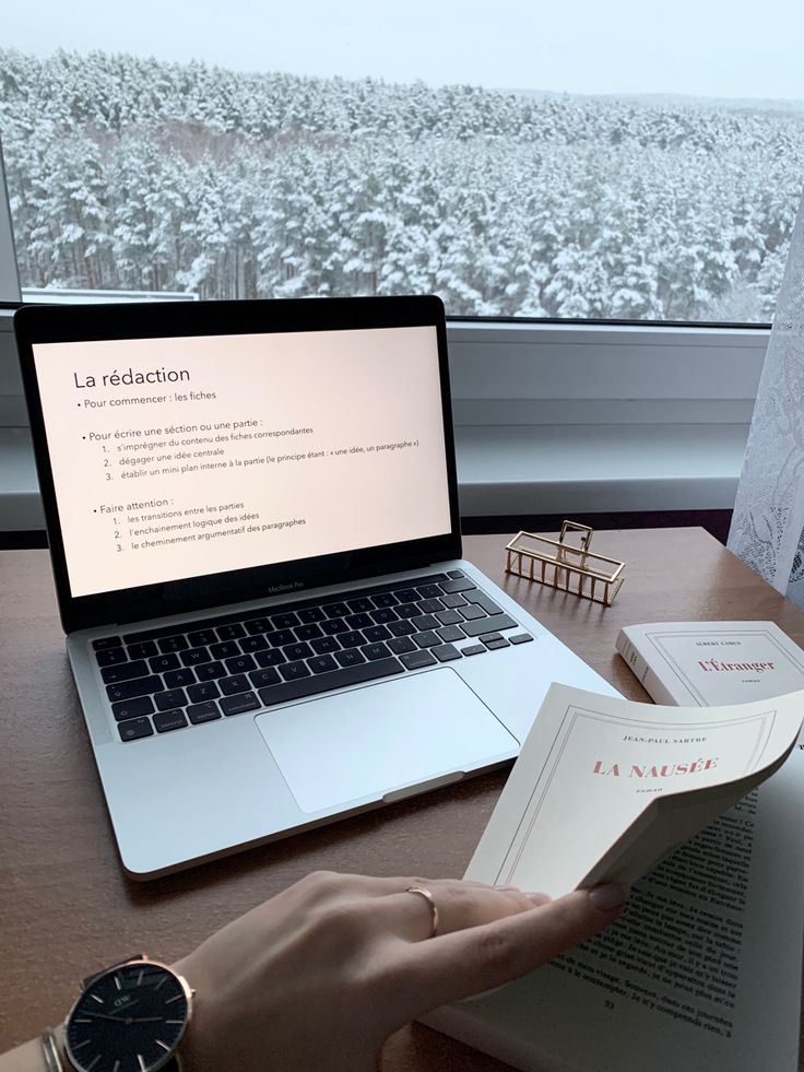 a person is reading a book in front of a laptop on a desk next to a window