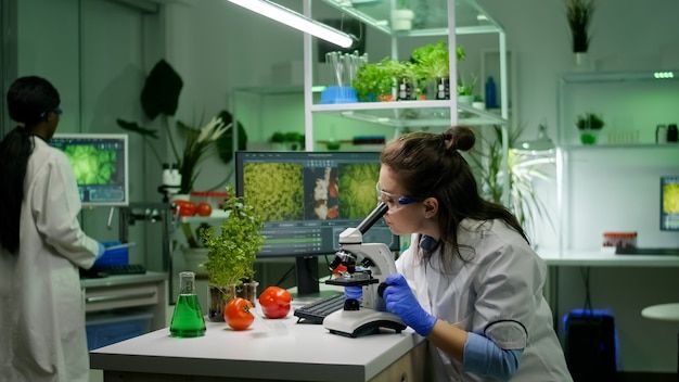 two women in white lab coats working with plants and microscopes on a table next to each other