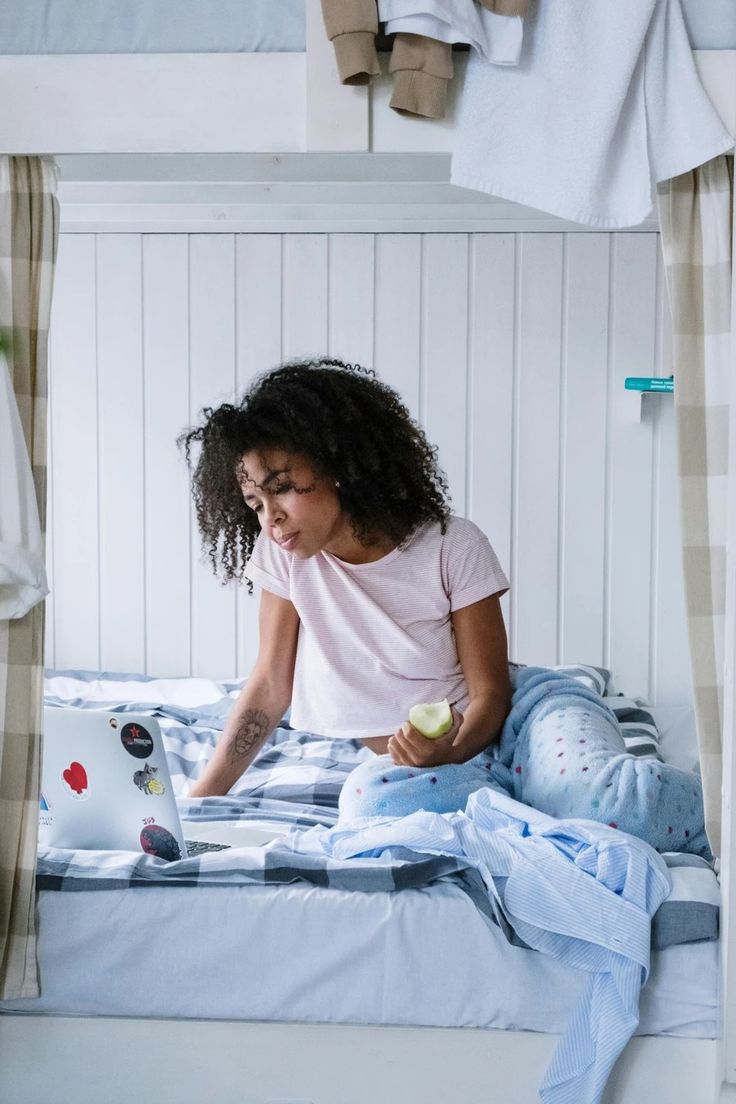 a woman sitting on top of a bed holding an apple and looking at her laptop