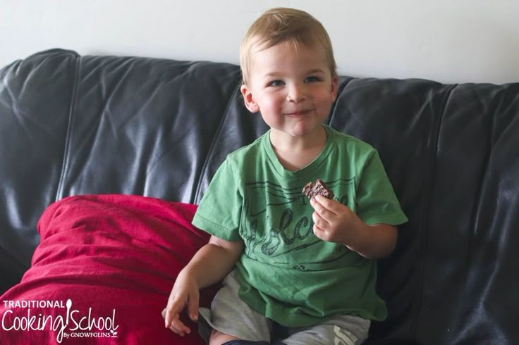 a young boy sitting on top of a black couch holding a piece of food in his hand