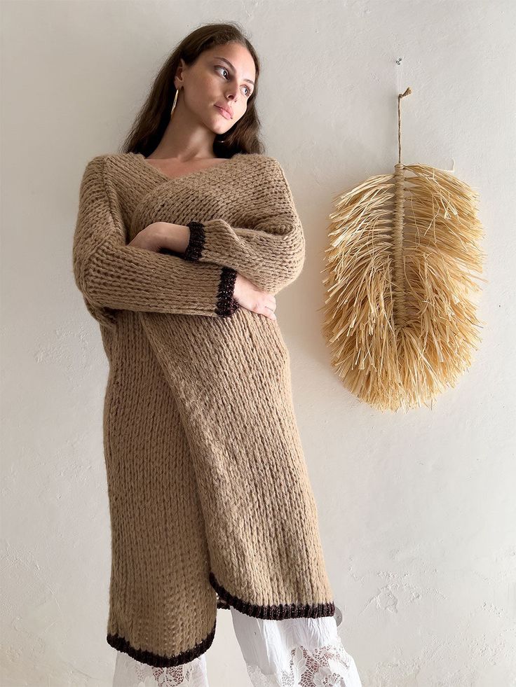 A person with long hair stands against a beige wall Long Beige Cozy Outerwear, Beige Long Cozy Outerwear, Long Brown Knitted Outerwear, Brown Knitted Long Outerwear, Long Brown Knitted Sweater Coat, Brown Long Knitted Sweater Coat, Oversized Beige Wool Sweater Coat, Beige Knitted Wool Sweater Coat, Beige Wool Knitted Sweater Coat