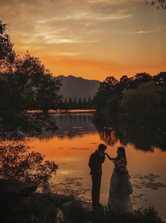 a bride and groom standing in front of a lake at sunset