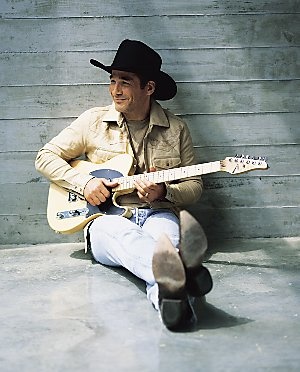 a man sitting on the ground with a guitar in his hand and wearing a cowboy hat