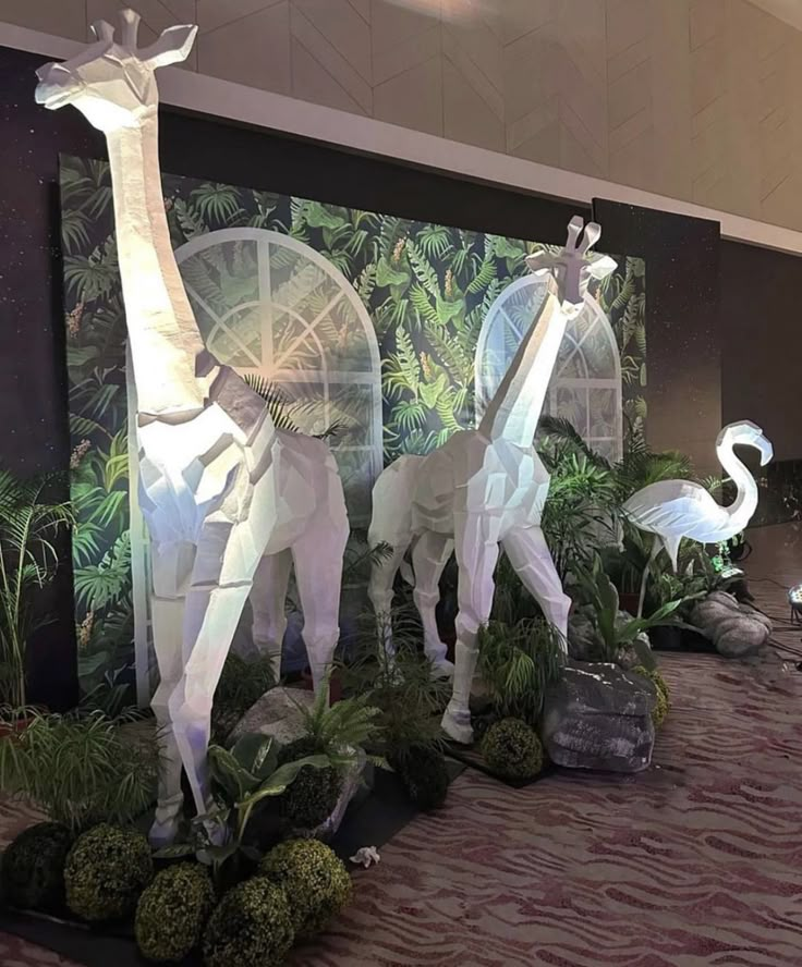 some statues of giraffes and flamingos in front of a wall with plants