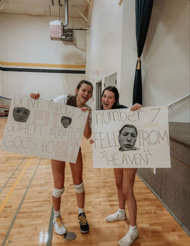 two girls holding up signs with faces on them in a gym floor, one girl is smiling and the other has her hands behind her head