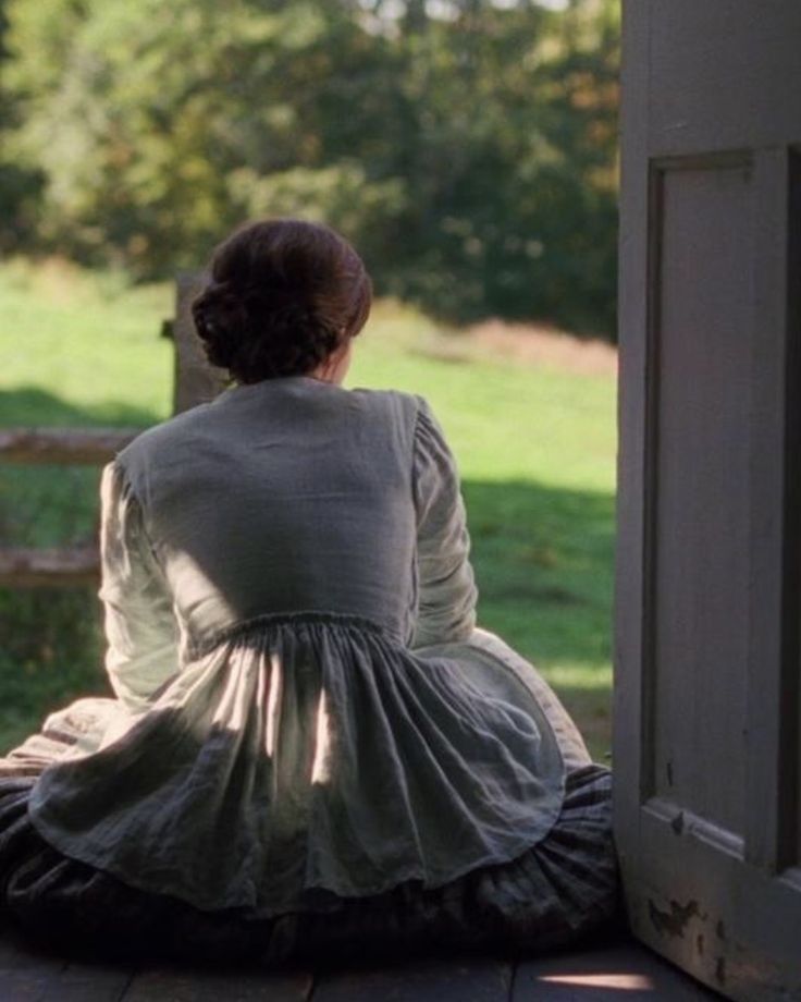 a woman sitting on top of a wooden floor next to an open door in front of a lush green field